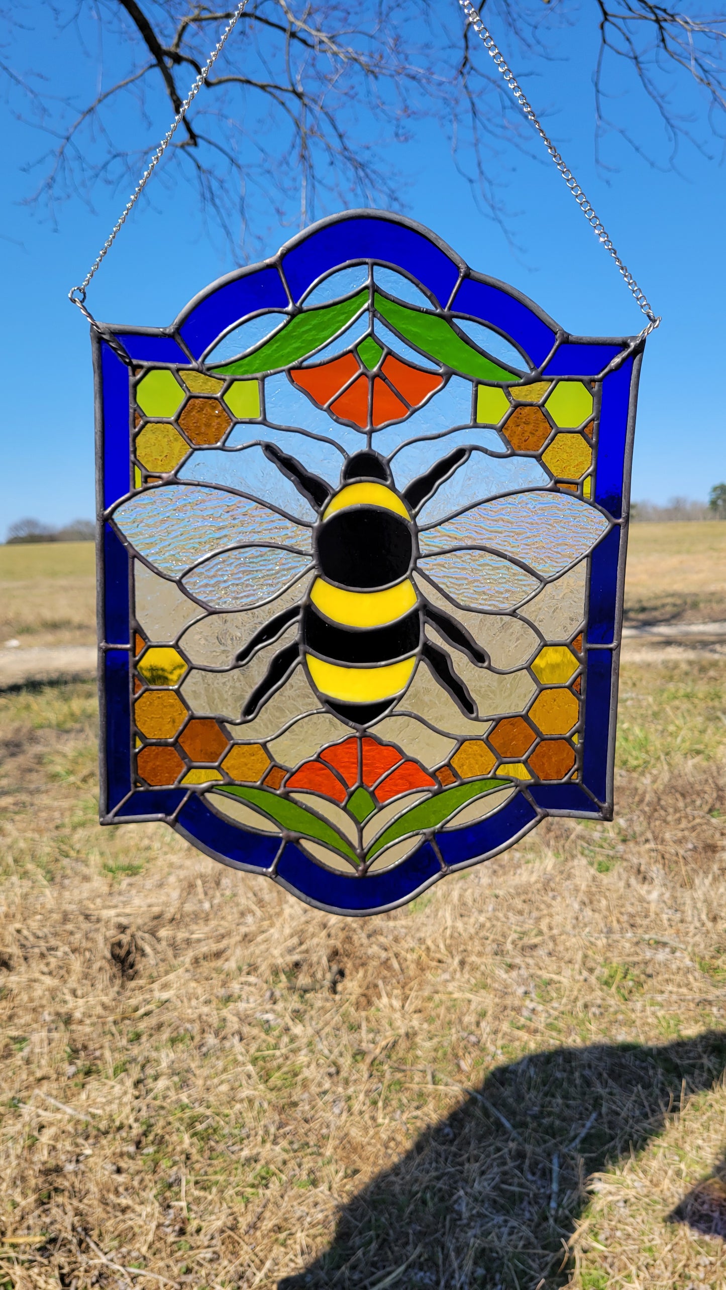 12x16 inch honeybee stained glass window panel featuring blue border, honeycomb pattern, and central bee hanging outdoors in sunlight.