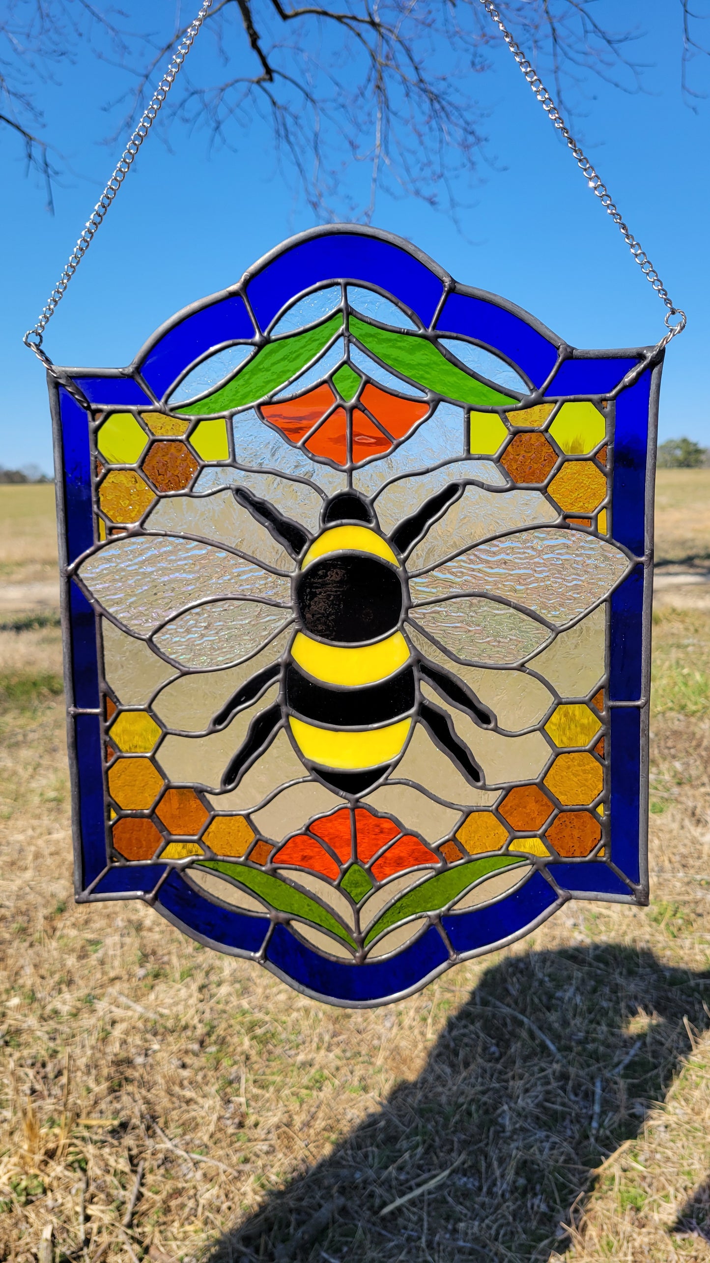 Stained glass honeybee suncatcher with honeycomb details and floral accents, hanging from a chain against a blue sky background.