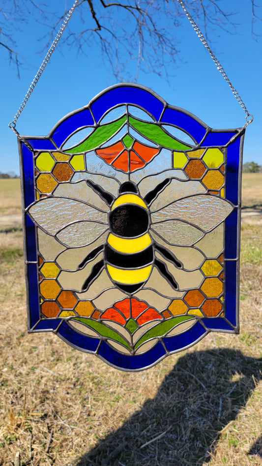 Stained glass honeybee suncatcher with honeycomb details and floral accents, hanging from a chain against a blue sky background.